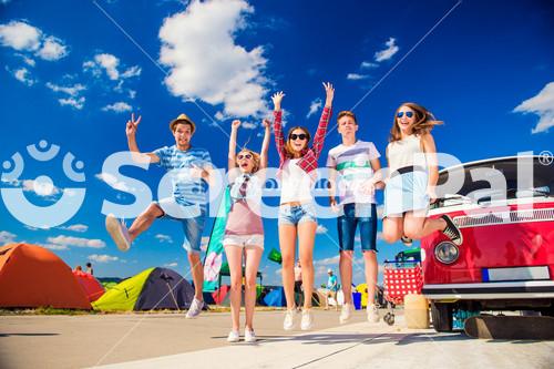 Group Of Teenage Boys And Girls At Summer Music Festival Jumping By Vintage Red Campervan